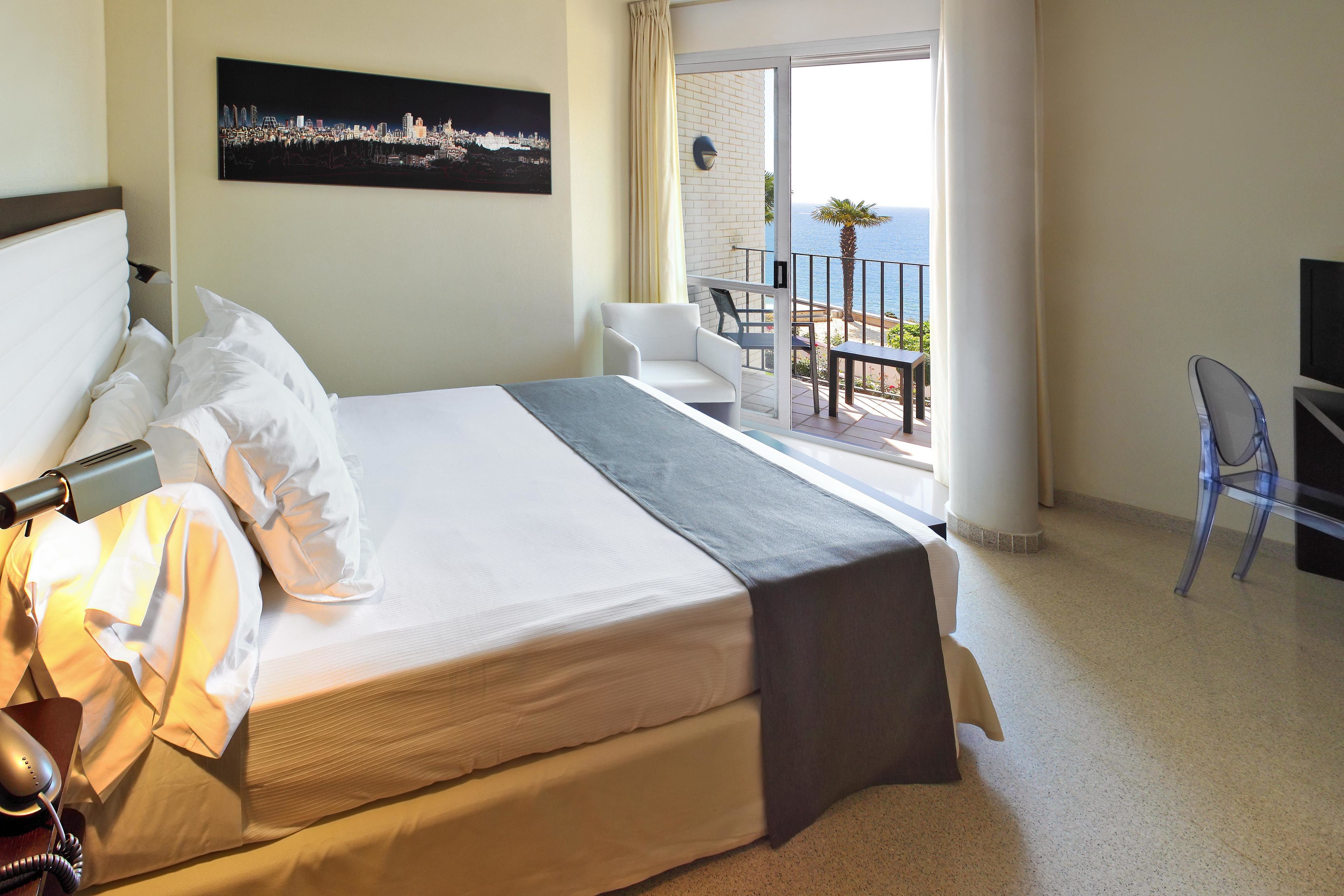 Una camera d'albergo con un letto, una scrivania, una sedia, una televisione e un balcone vista mare.