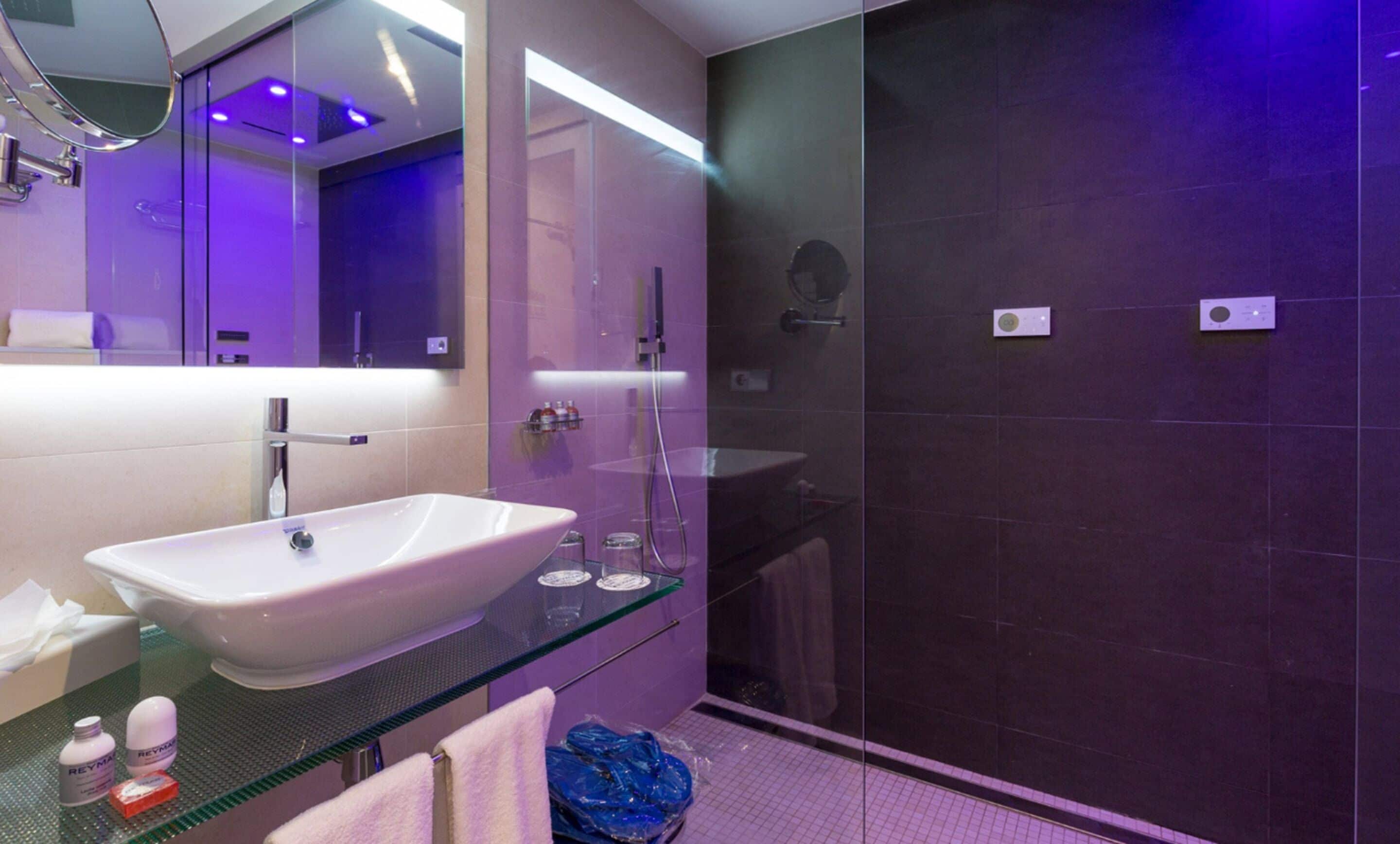 A modern bathroom with a large white sink, a glass countertop, and a dark tiled shower area.