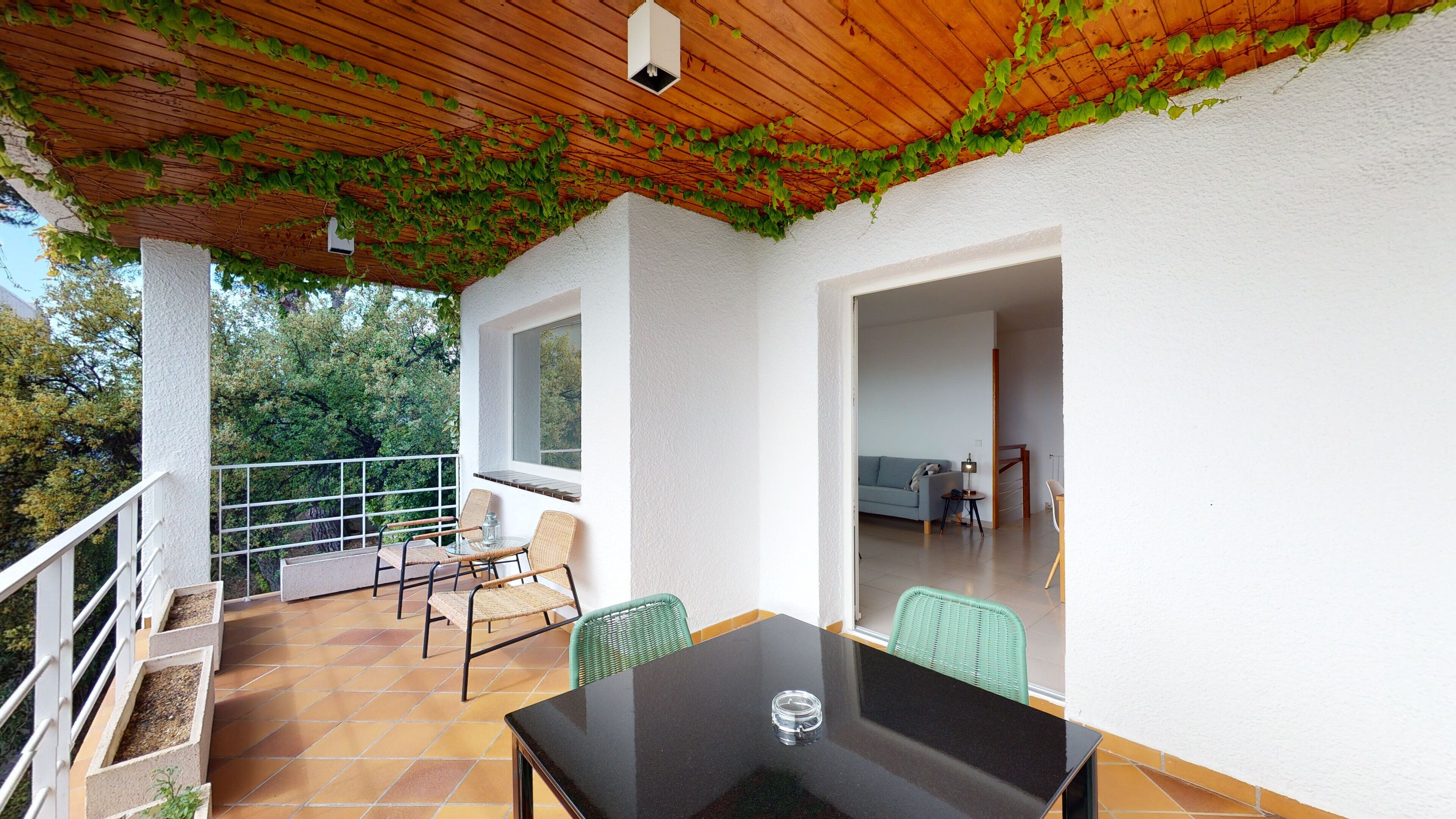 A covered patio with a table and chairs, a view of the outdoors, and a white wall with a window.