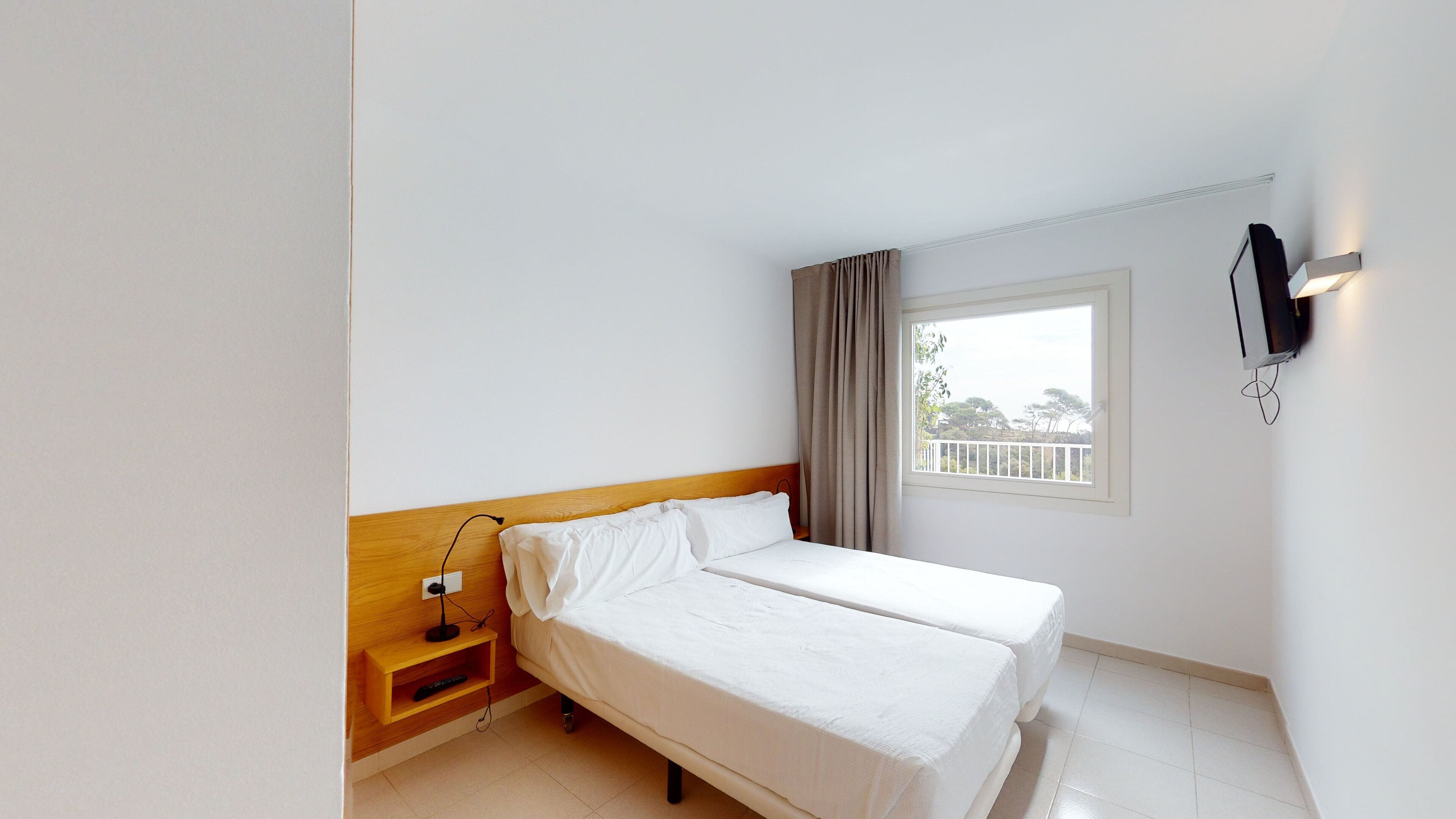 A hotel room with a single bed, a bedside table with a lamp, a TV mounted on the wall, and a window with a view of trees and a balcony.