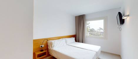 A hotel room with a single bed, a bedside table with a lamp, a TV mounted on the wall, and a window with a view of trees and a balcony.