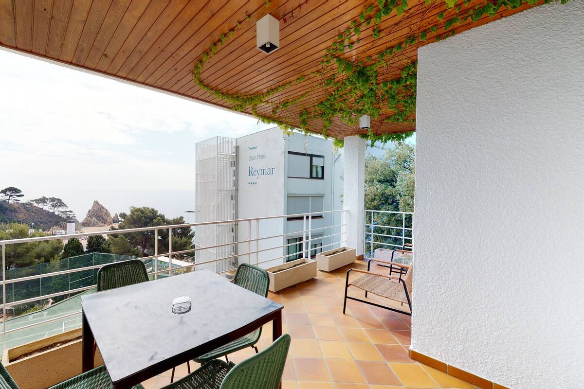 A balcony with a table and chairs, overlooking a scenic view of a building labeled "Premier Gran Hotel Reymar" and a coastal landscape.