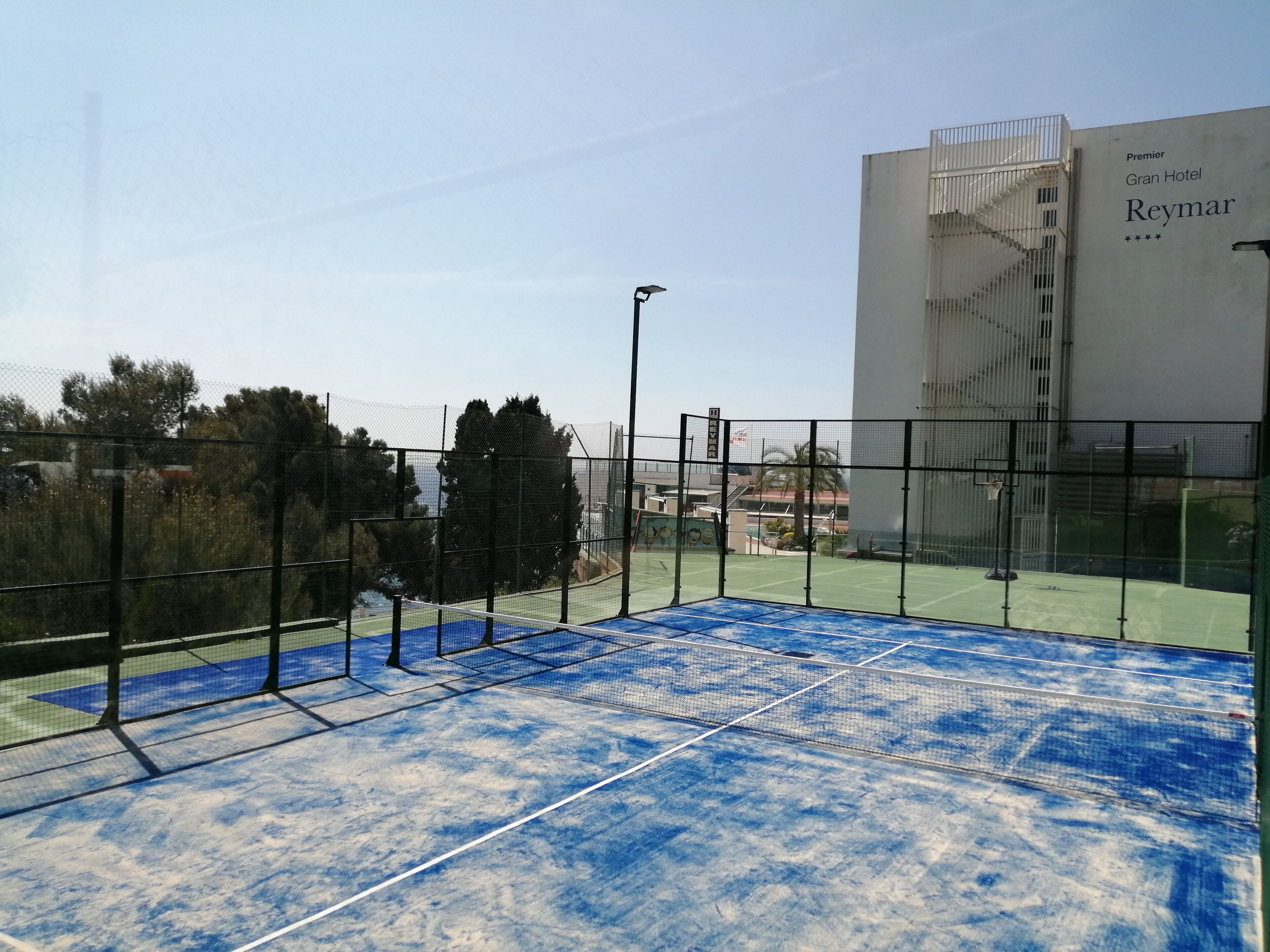 A tennis court with a net, surrounded by a fence, and a building labeled "Premier Gran Hotel Reymar" in the background.