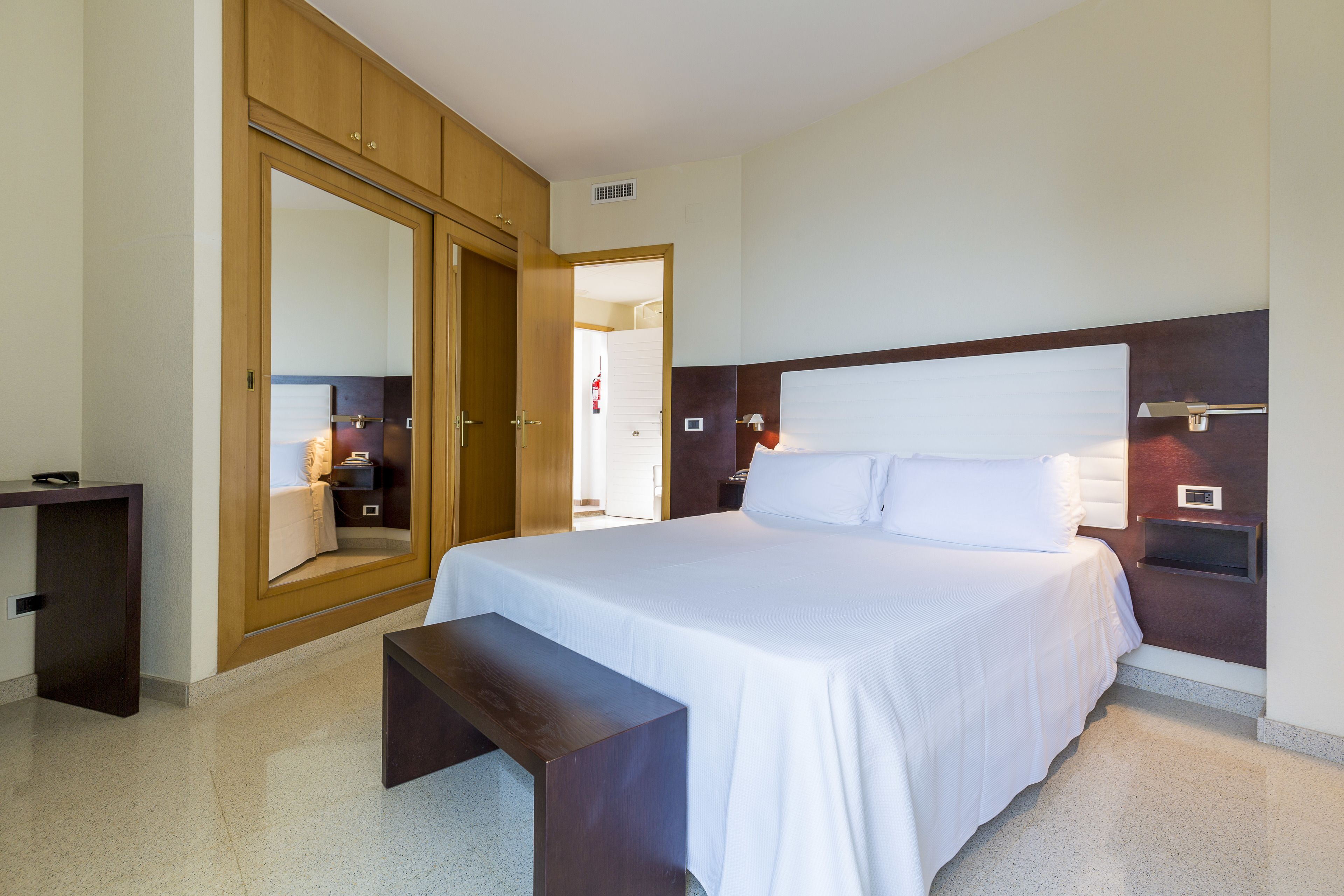 A hotel room with a large bed, a wooden wardrobe, a bedside table, and a bench.