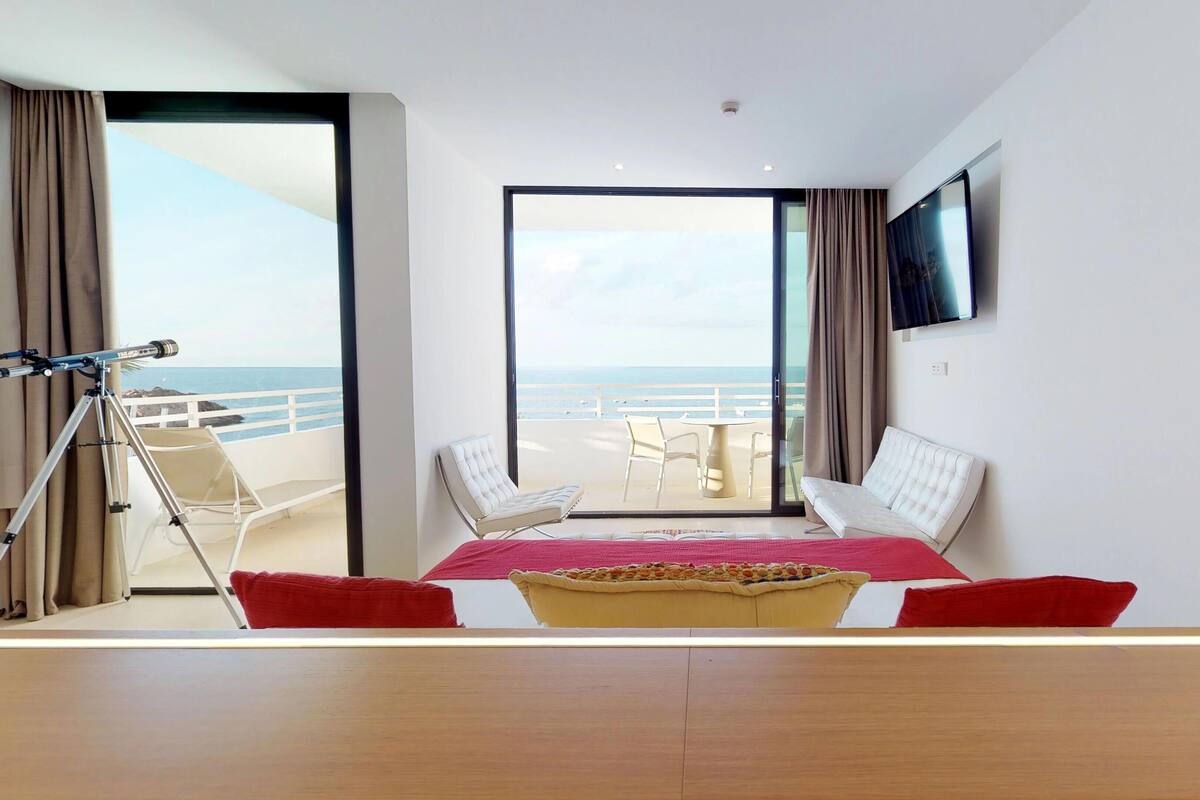 A modern hotel room with a large balcony, a bed, a TV, and a telescope.