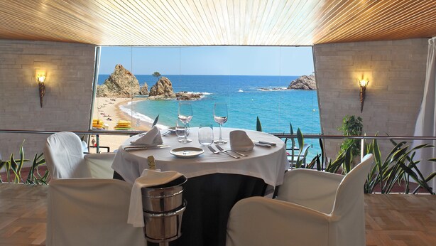 A dining area with a table set for two, overlooking a beach and the sea. - Gran Hotel Reymar (Tossa de Mar)