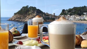 A table with breakfast items and a view of the sea and a coastal town. - Gran Hotel Reymar (Tossa de Mar)