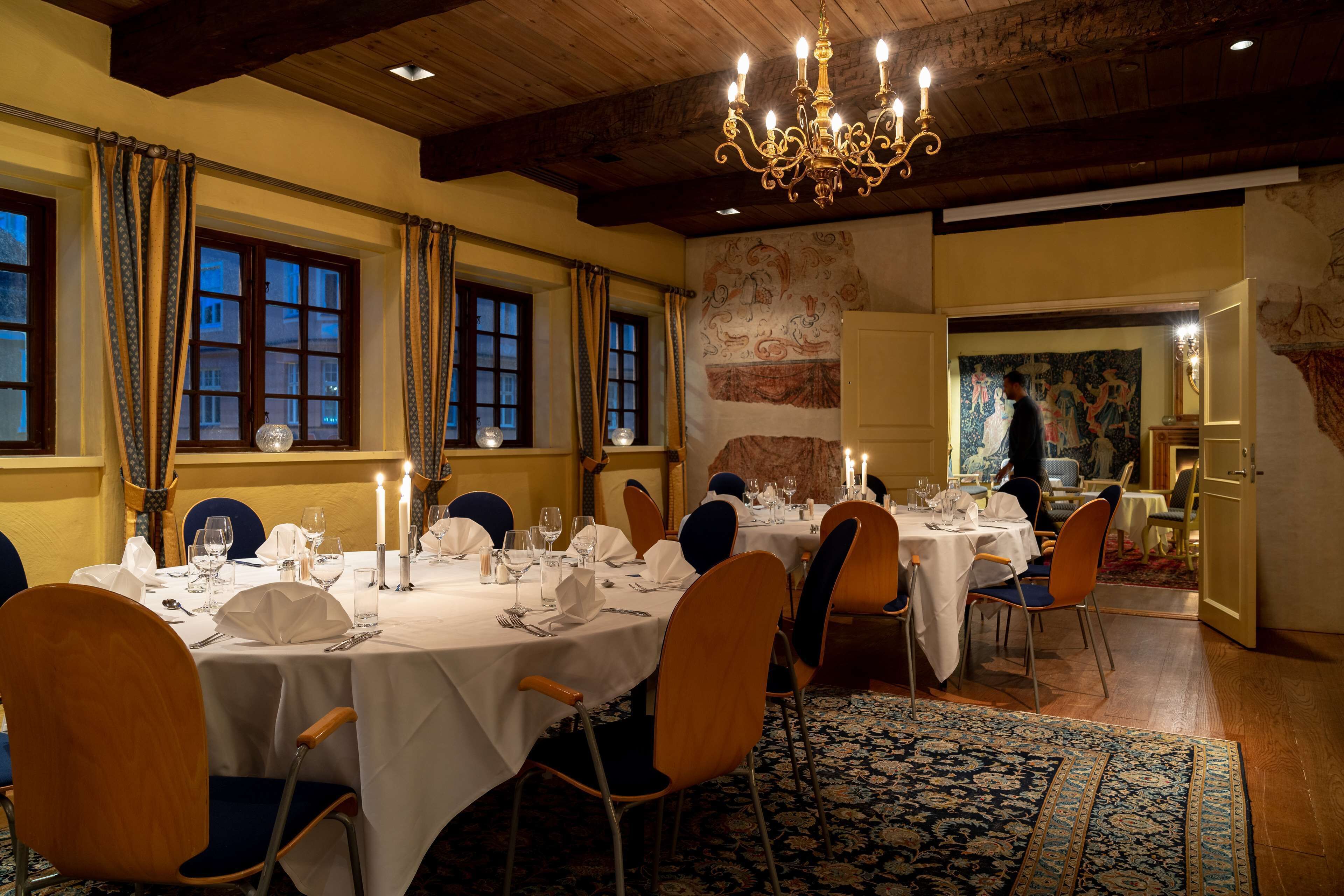 Uma sala de jantar com mesas redondas dispostas para refeição, com toalhas brancas, velas e cadeiras.