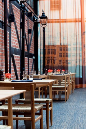 A row of wooden tables and chairs in a restaurant with brick walls and a large window.