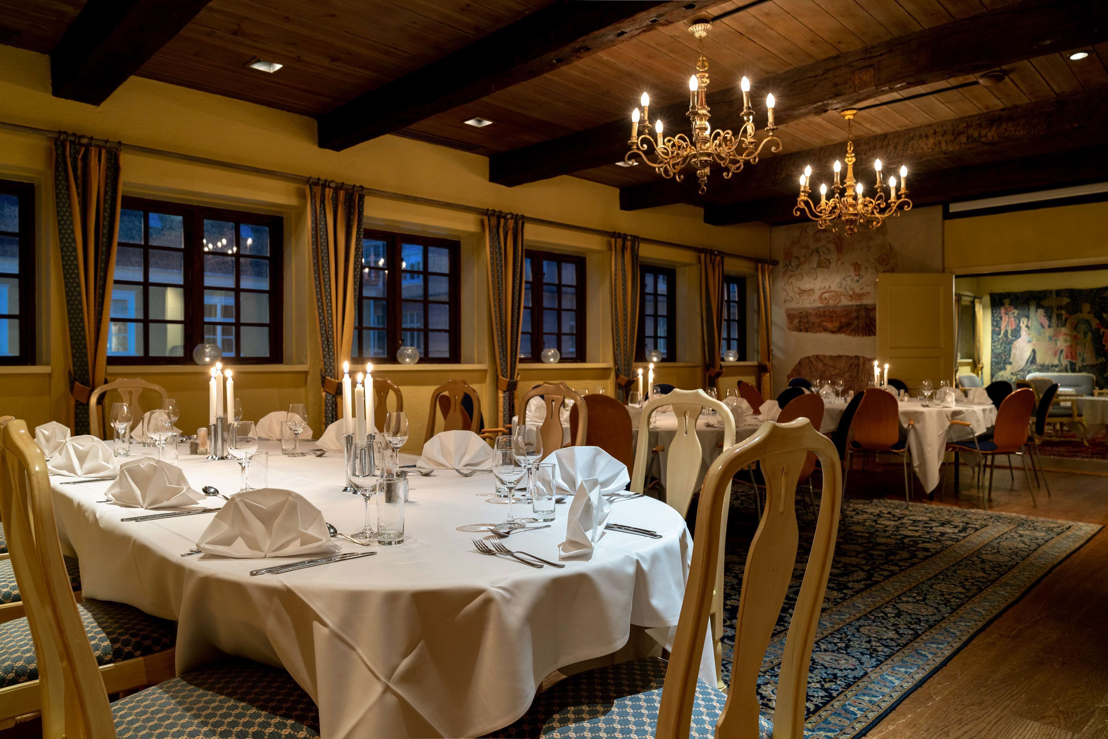Une salle à manger élégante, avec des tables rondes dressées pour le repas, ornées de nappes blanches, de bougies et de porcelaine de qualité.