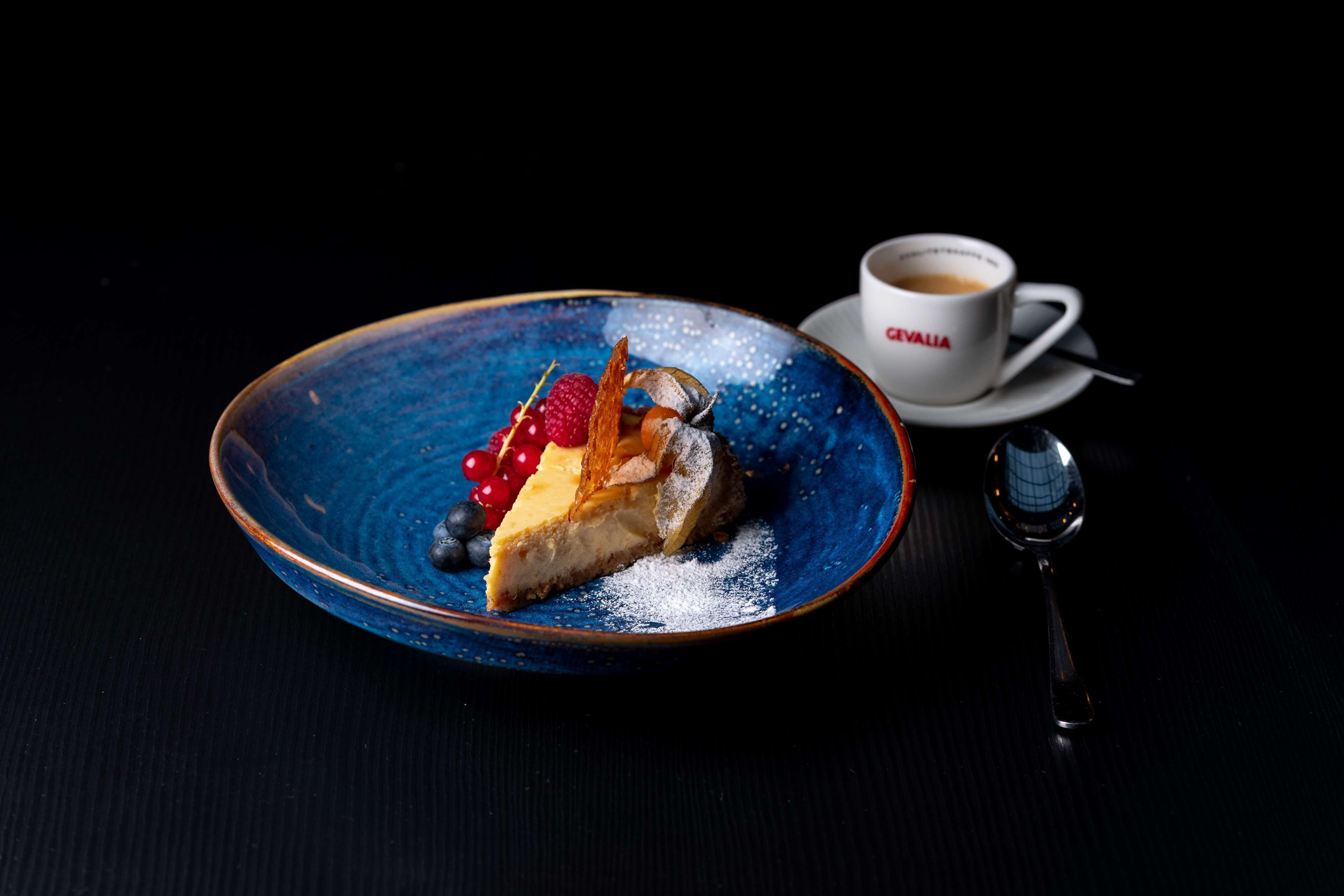 Ein Dessertteller mit einem Stück Käsekuchen, Beeren und einer Dekoration, zusammen mit einer Tasse Kaffee auf einem Untertasse.