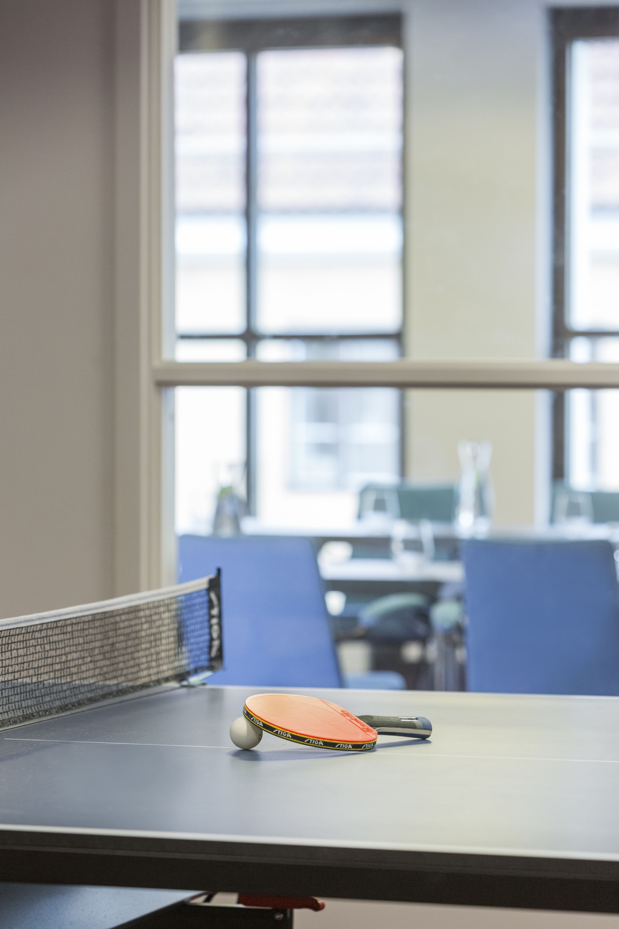 A table tennis table with a paddle and ball, a window with blinds, and a blurred office background.