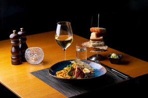 Una mesa de madera con un plato azul de pasta, una copa de vino blanco, un vaso de agua, saleros y pimenteros, un bol pequeño de verduras, y una pila de pan.