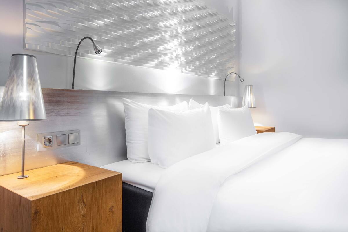 A modern hotel room with a large bed, white bedding, and a unique textured wall above the bed. There are two bedside tables with lamps.