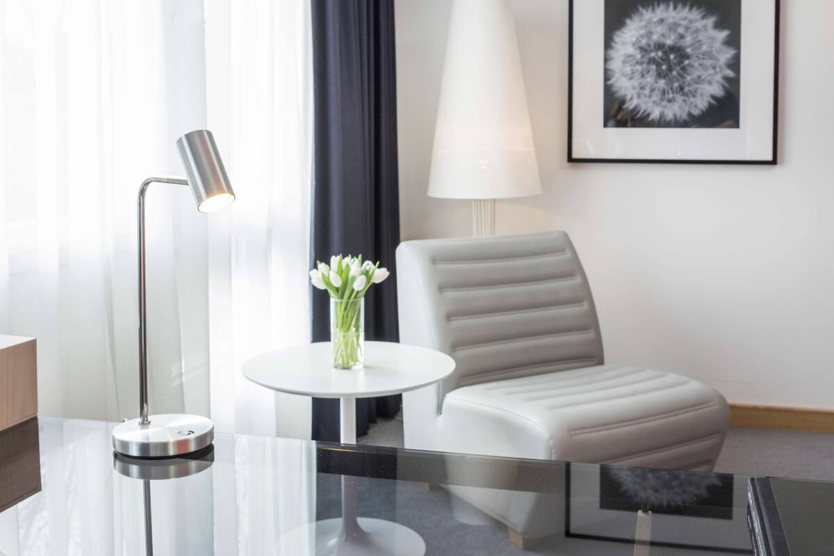 A modern hotel room with a glass table, a white chair, a lamp, and a framed picture on the wall.