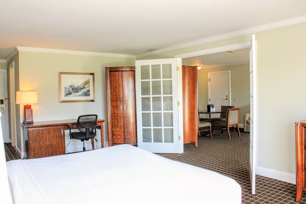 A hotel room with a bed, desk, chair, and a door leading to a dining area.