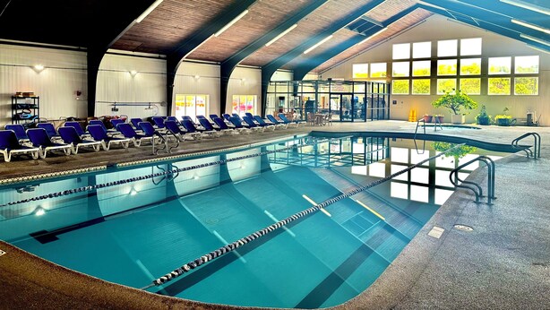 An indoor swimming pool with clear lane dividers, a lounge area with blue chairs, and a high ceiling with wooden beams. - Cape Cod Irish Village (Hyannis)