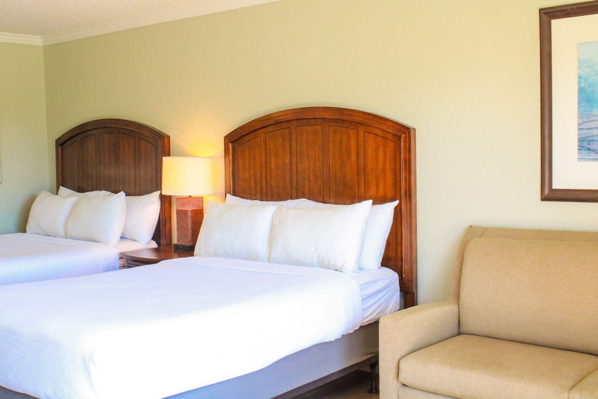 A hotel room with two beds, a nightstand with a lamp, and a beige armchair.