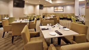 A hotel dining area with multiple tables and chairs, a flat-screen TV mounted on the wall, and a cityscape painting on the wall.