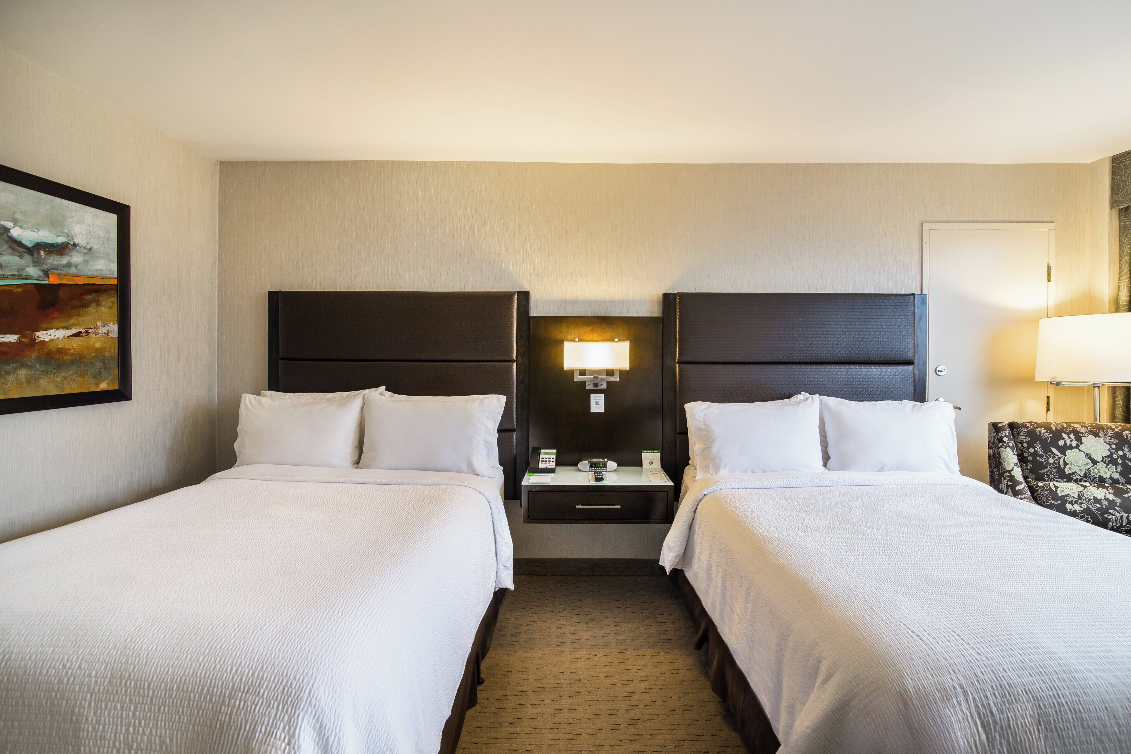 A hotel room with two beds, a nightstand, a lamp, and a painting on the wall.