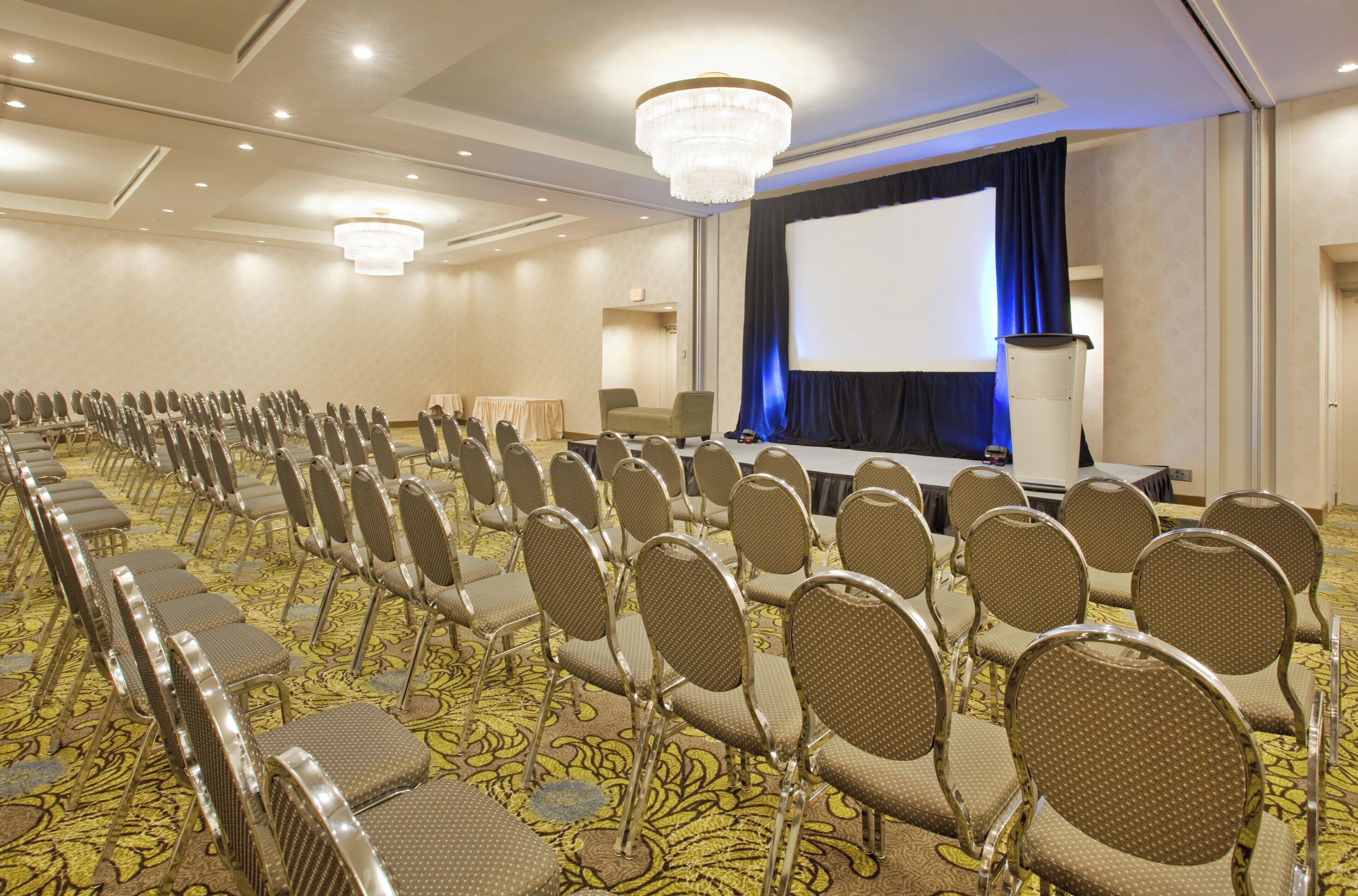A conference room with rows of chairs, a stage with a screen, and a chandelier.