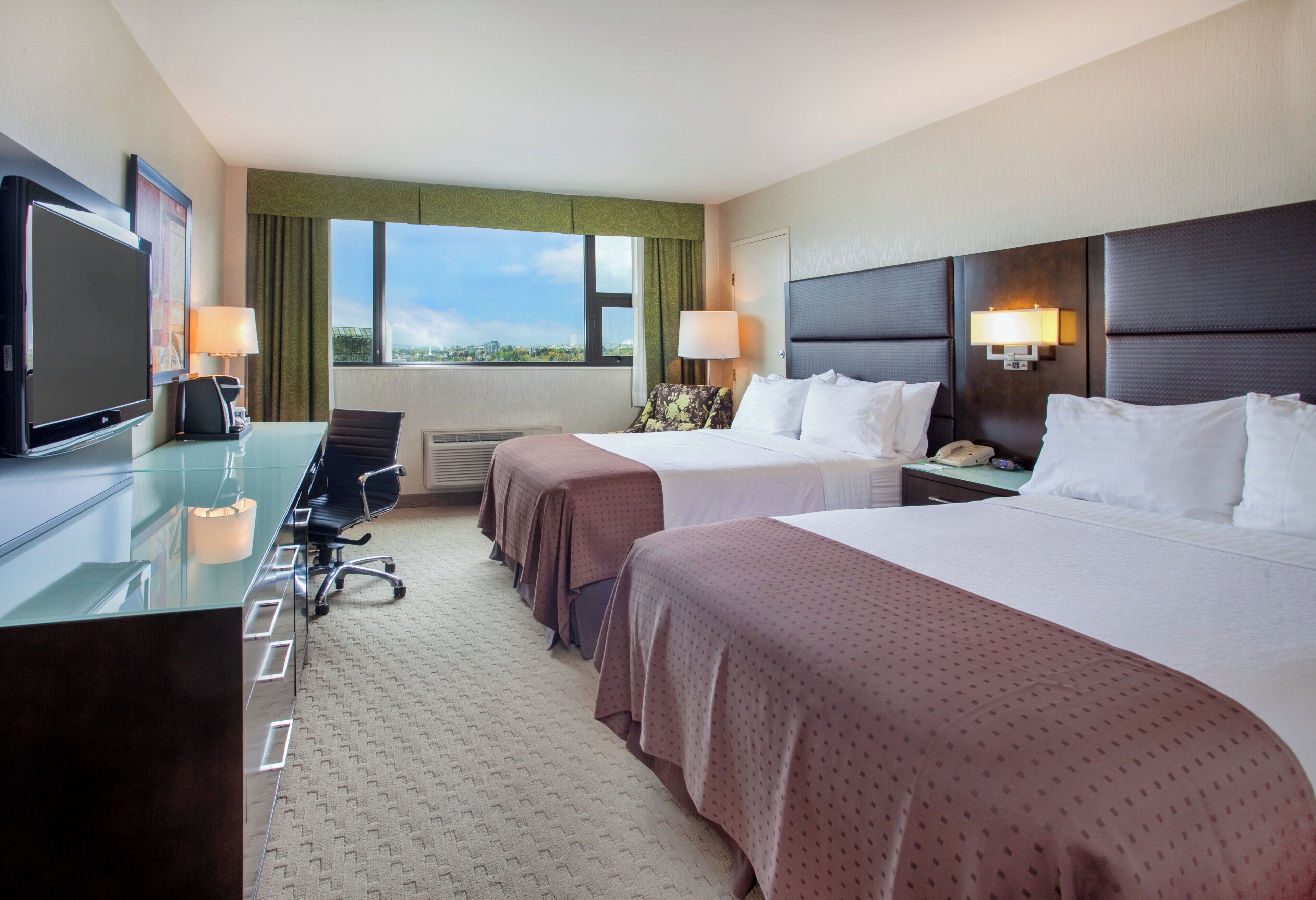 A hotel room with two beds, a desk with a flat-screen TV, a chair, and a window with a view of the city.