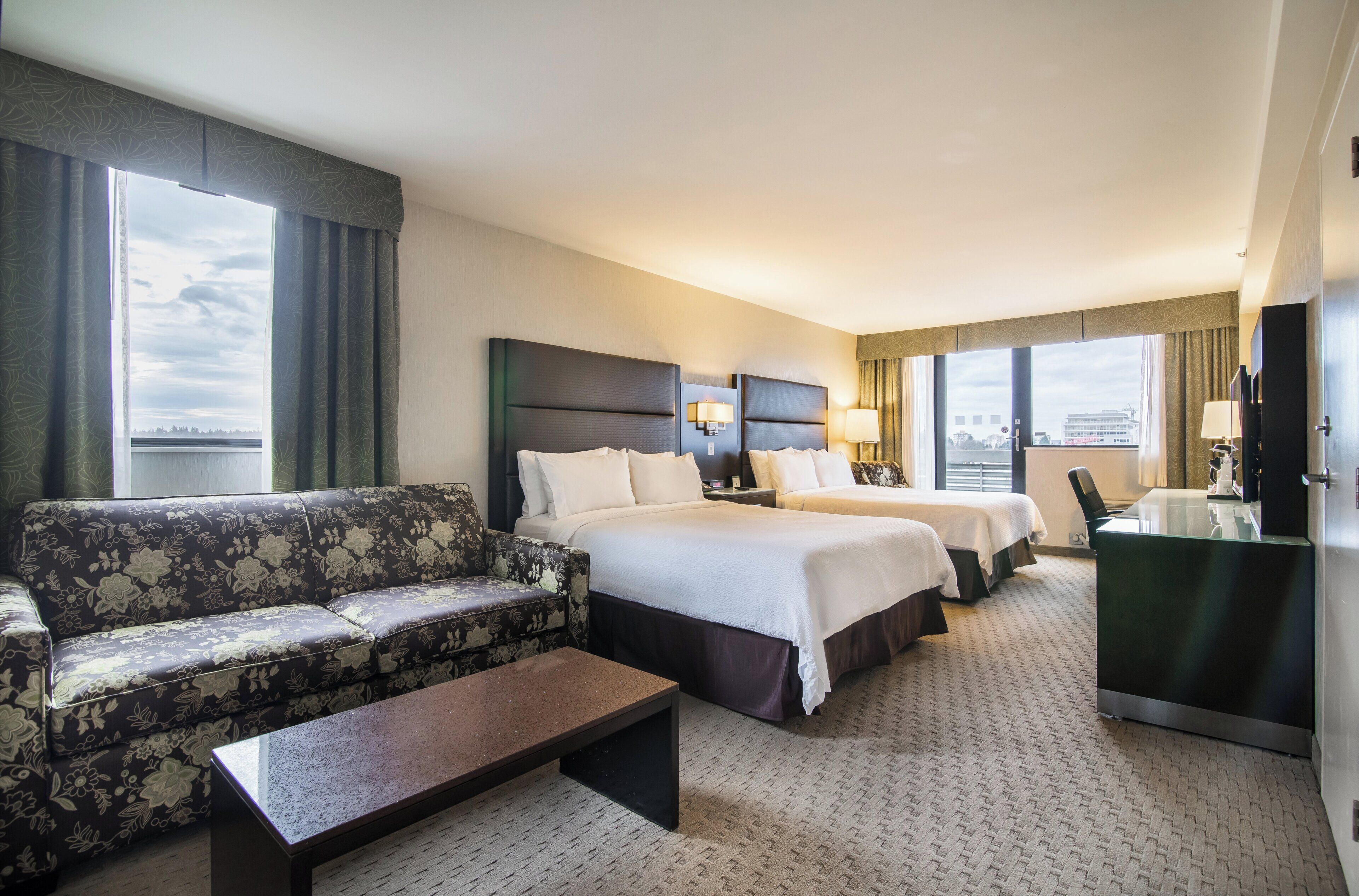 A hotel room with a sofa, two beds, a desk, and a view of the city.