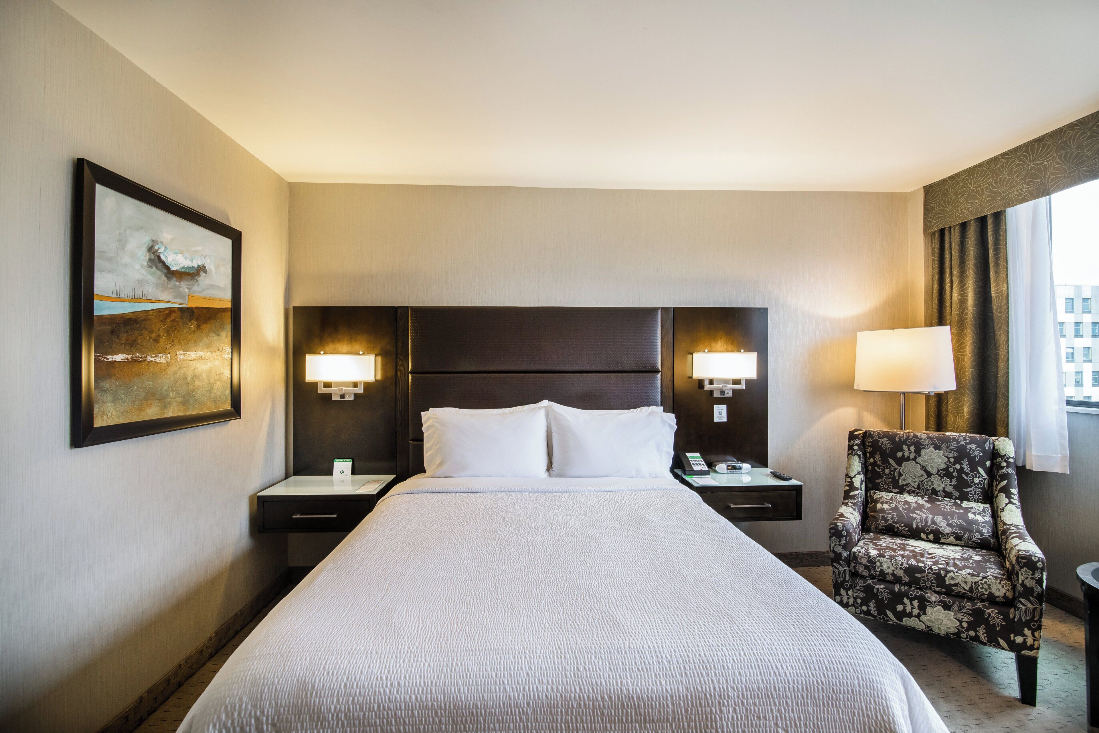 A hotel room with a large bed, two bedside tables, a chair, and a painting on the wall.