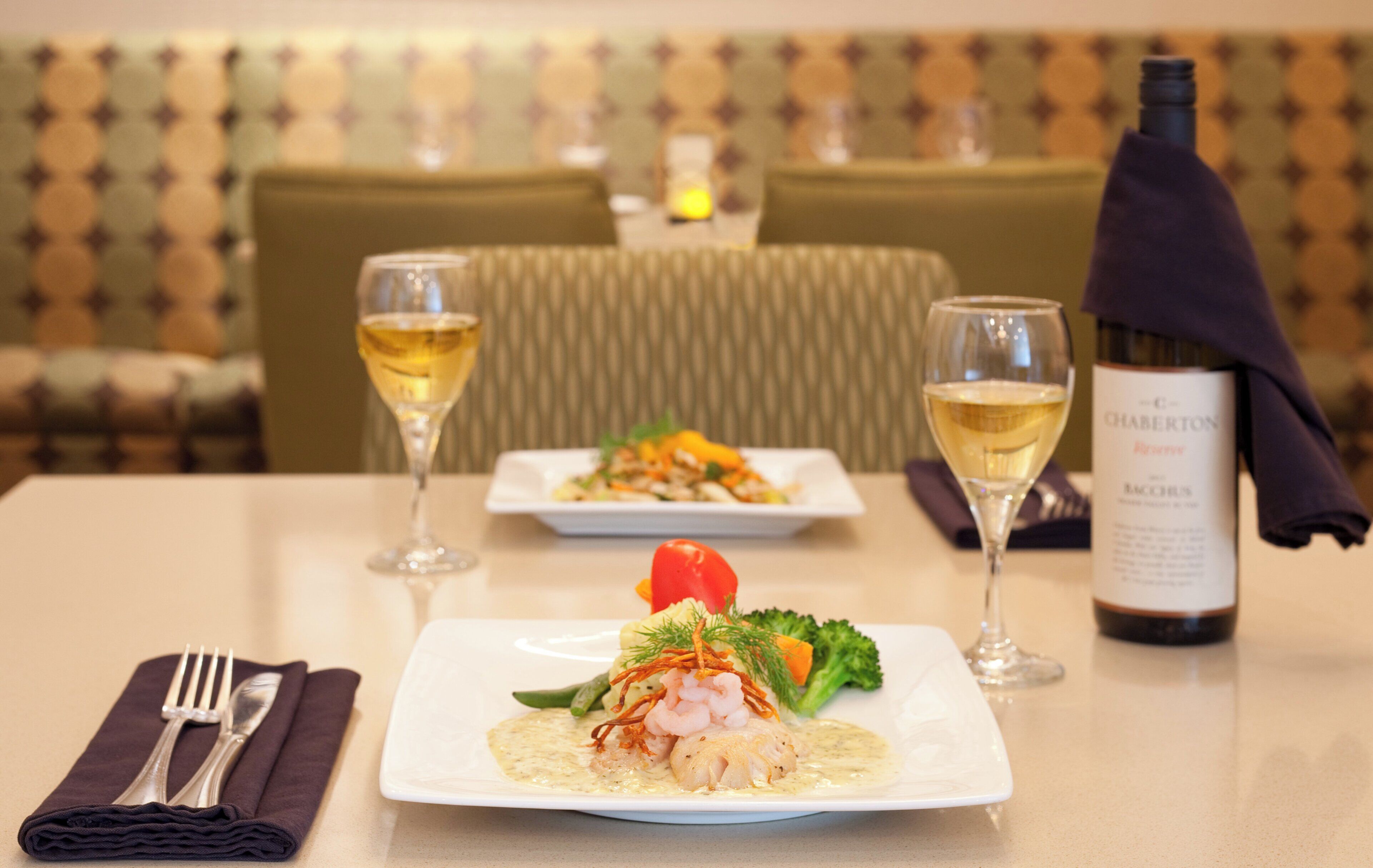 A dining table with two plates of food, two glasses of white wine, and a bottle of wine on a hotel table.