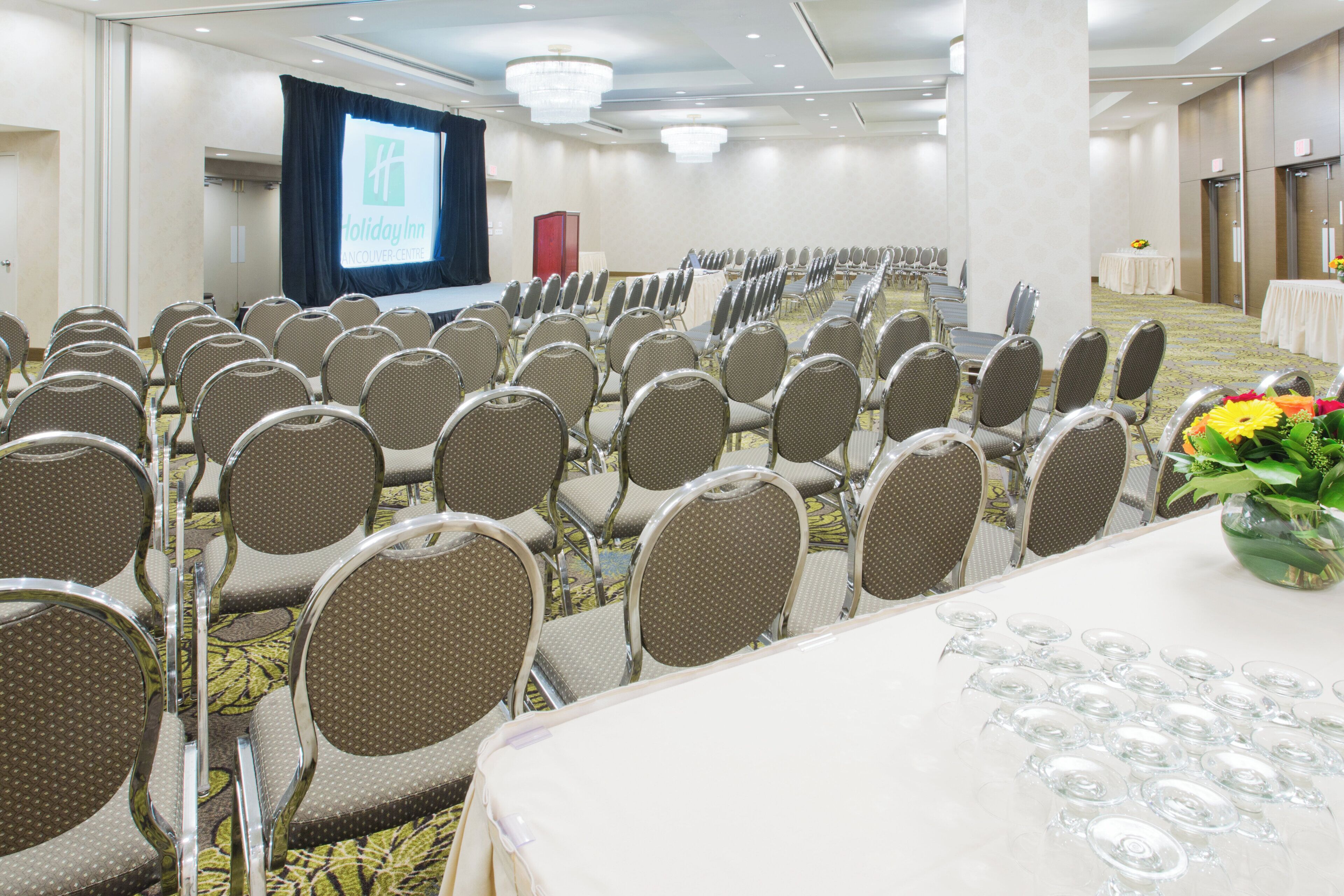 A conference room with rows of chairs, a stage with a screen, and a table with a vase of flowers.