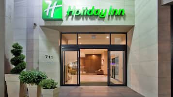 Entrada de un hotel Holiday Inn con puerta de vidrio, un letrero con el número 711 y plantas en macetas a la izquierda.