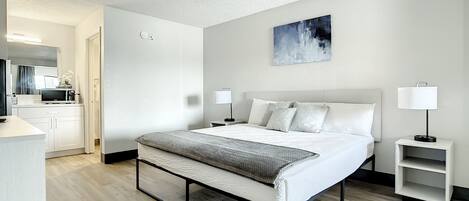 Ett modernt hotellrum med en stor säng, sänglampor, ett litet nattduksbord och utsikt över köksdelen.