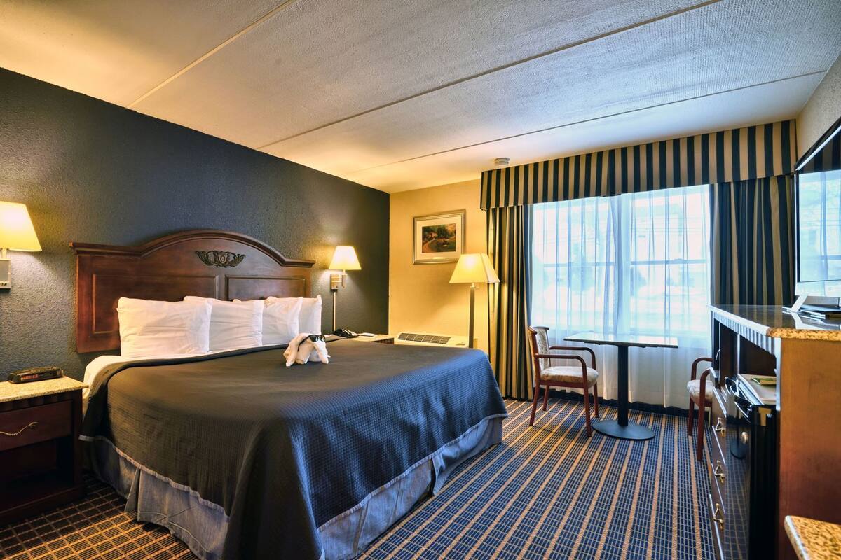 A hotel room with a large bed, bedside lamps, a desk with a chair, a window with curtains, and a painting on the wall.
