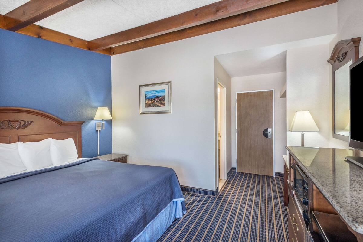 A hotel room with a large bed, a wooden headboard, a picture on the wall, a door leading to another room, and a bathroom with a mirror and sink.