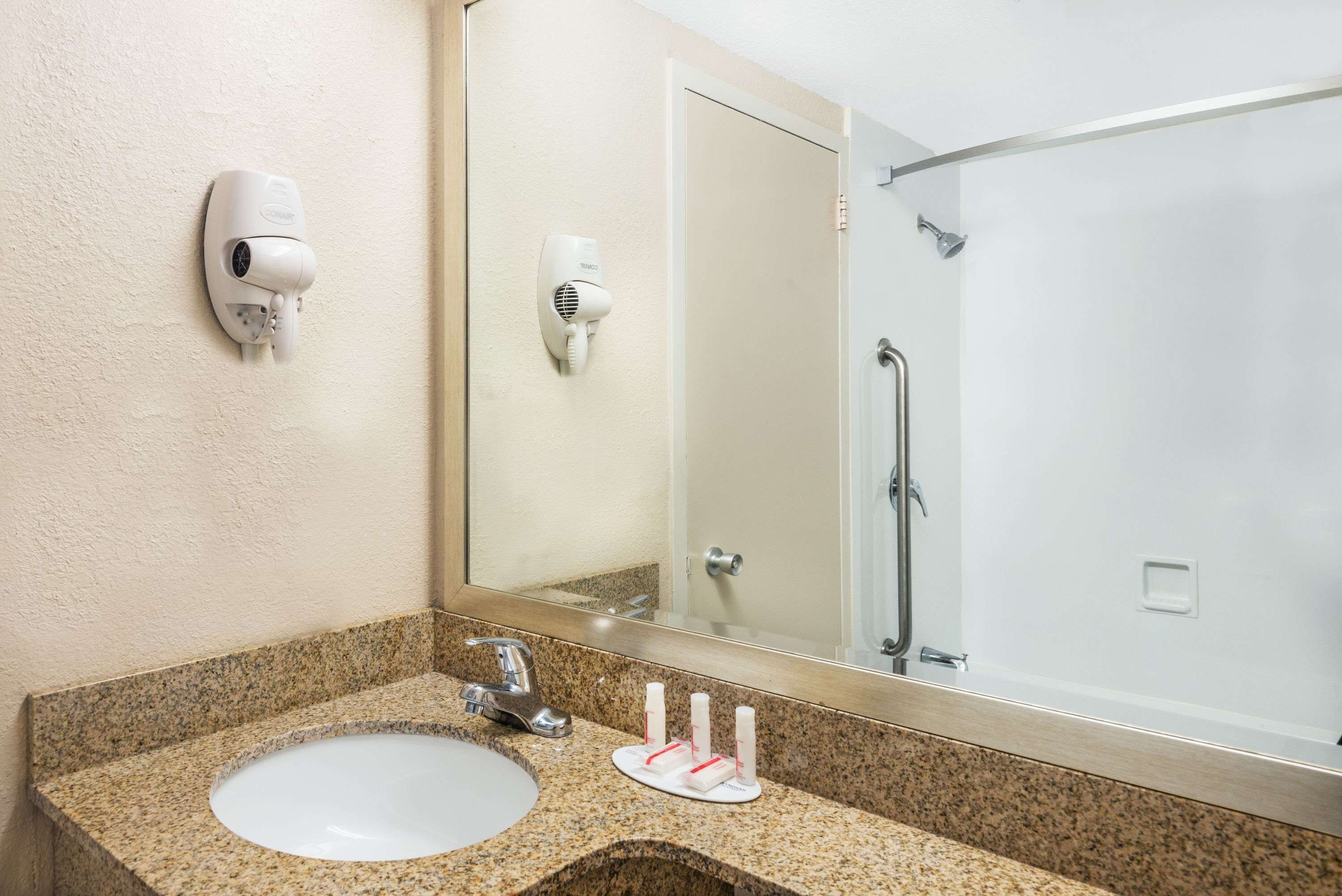 A bathroom with a granite countertop, a white sink, a mirror, a hair dryer, and a shower.