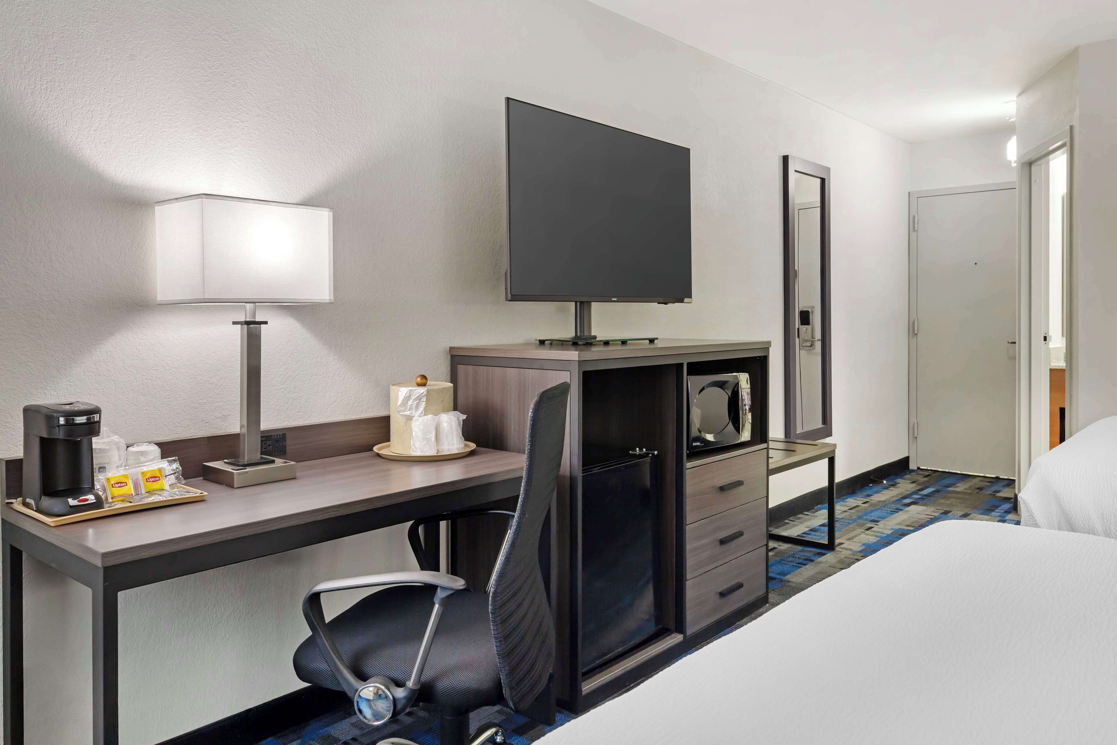 A hotel room with a bed, a desk with a lamp, a television, a chair, a coffee maker, and a minibar.