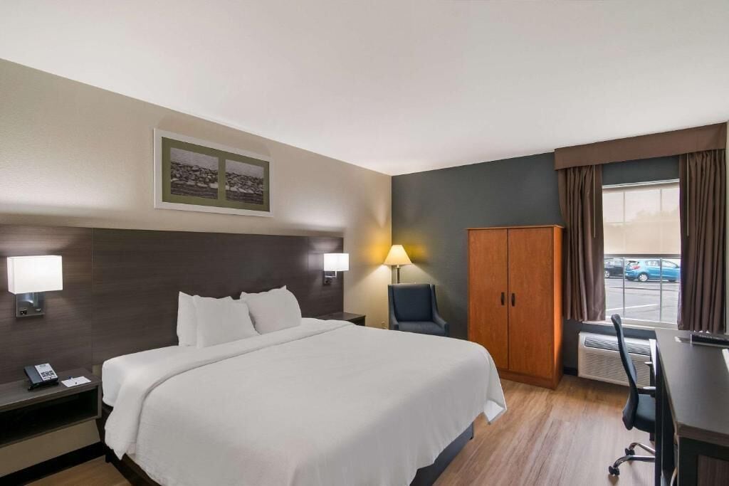 Habitación de hotel con cama, lámparas de noche, escritorio, silla, armario y ventana con cortinas.