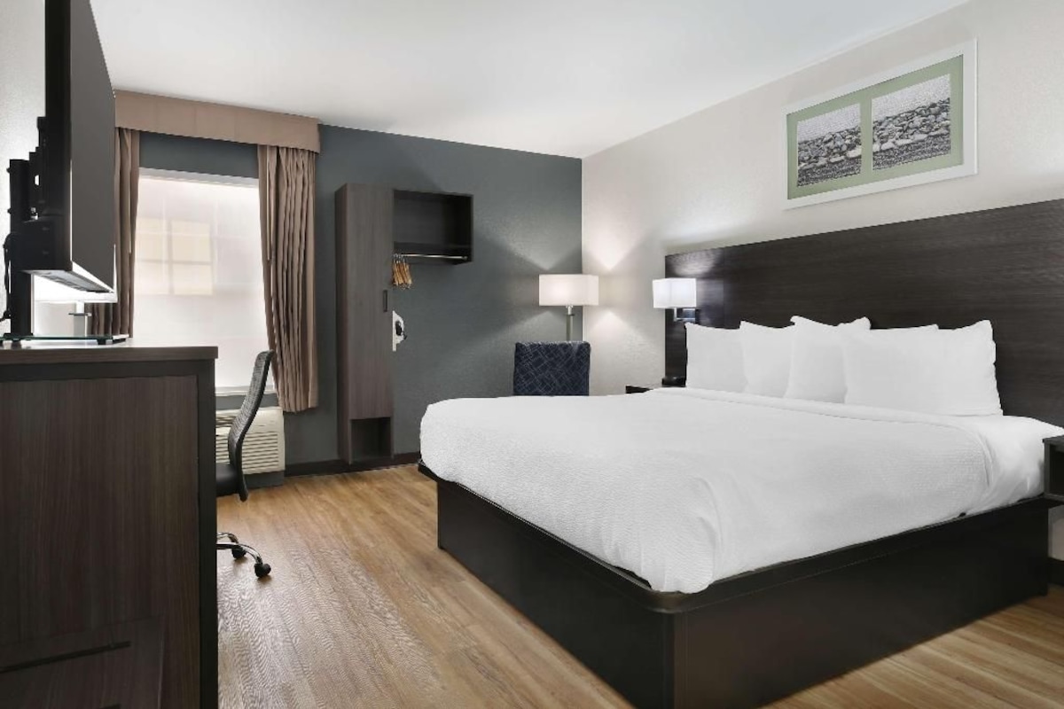 Habitación de hotel con una cama grande, un escritorio con lámpara, una silla y un televisor en la pared.