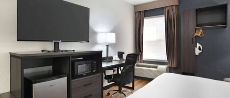 Ein Hotelzimmer mit einem Bett, einem Schreibtisch mit Computer, einer Mikrowelle, einem Flachbildfernseher, einer Lampe und einem Fenster mit Vorhängen.