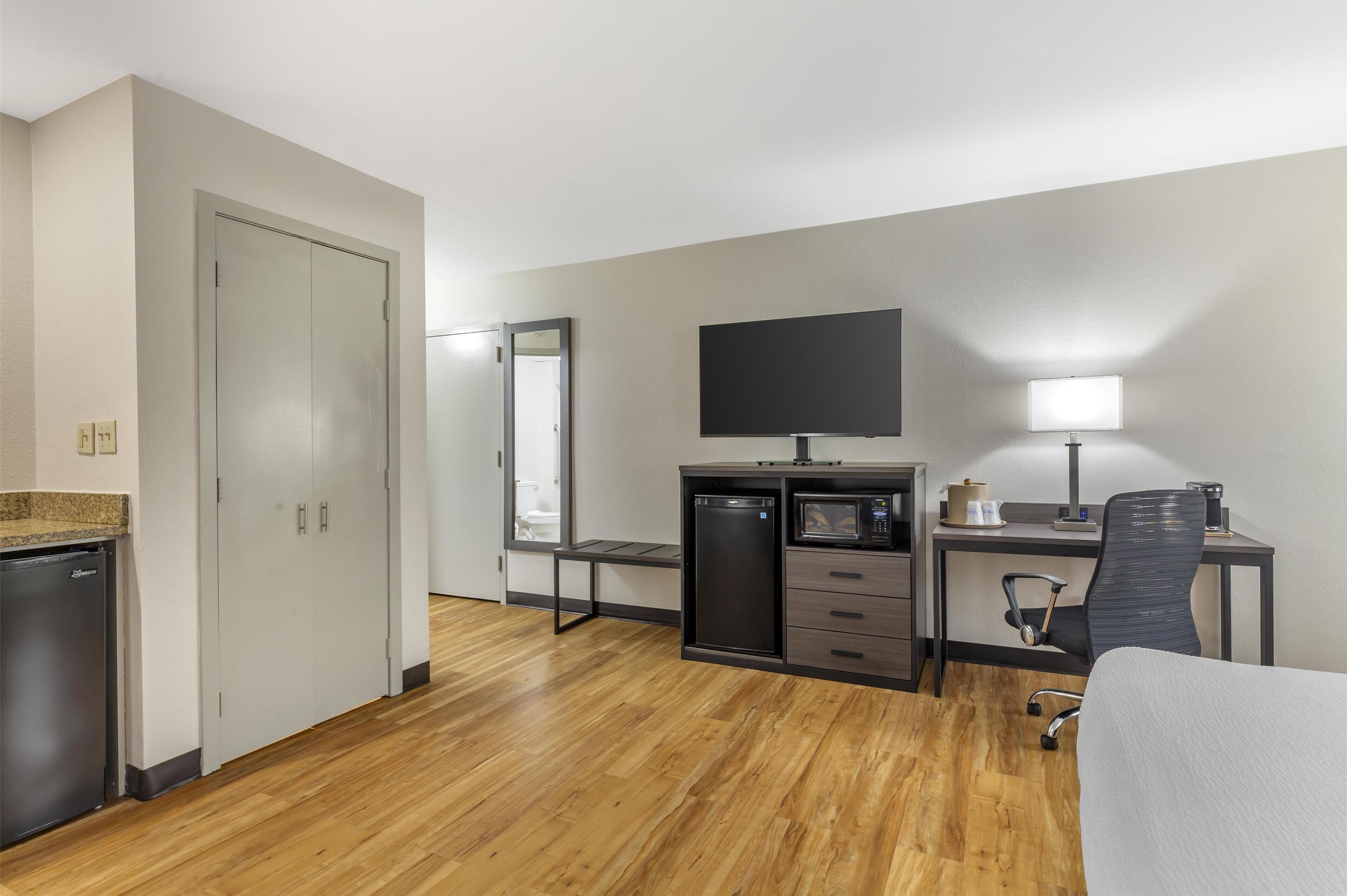A compact hotel room with a kitchenette, a bed, a desk with a computer and lamp, and a large mirror.