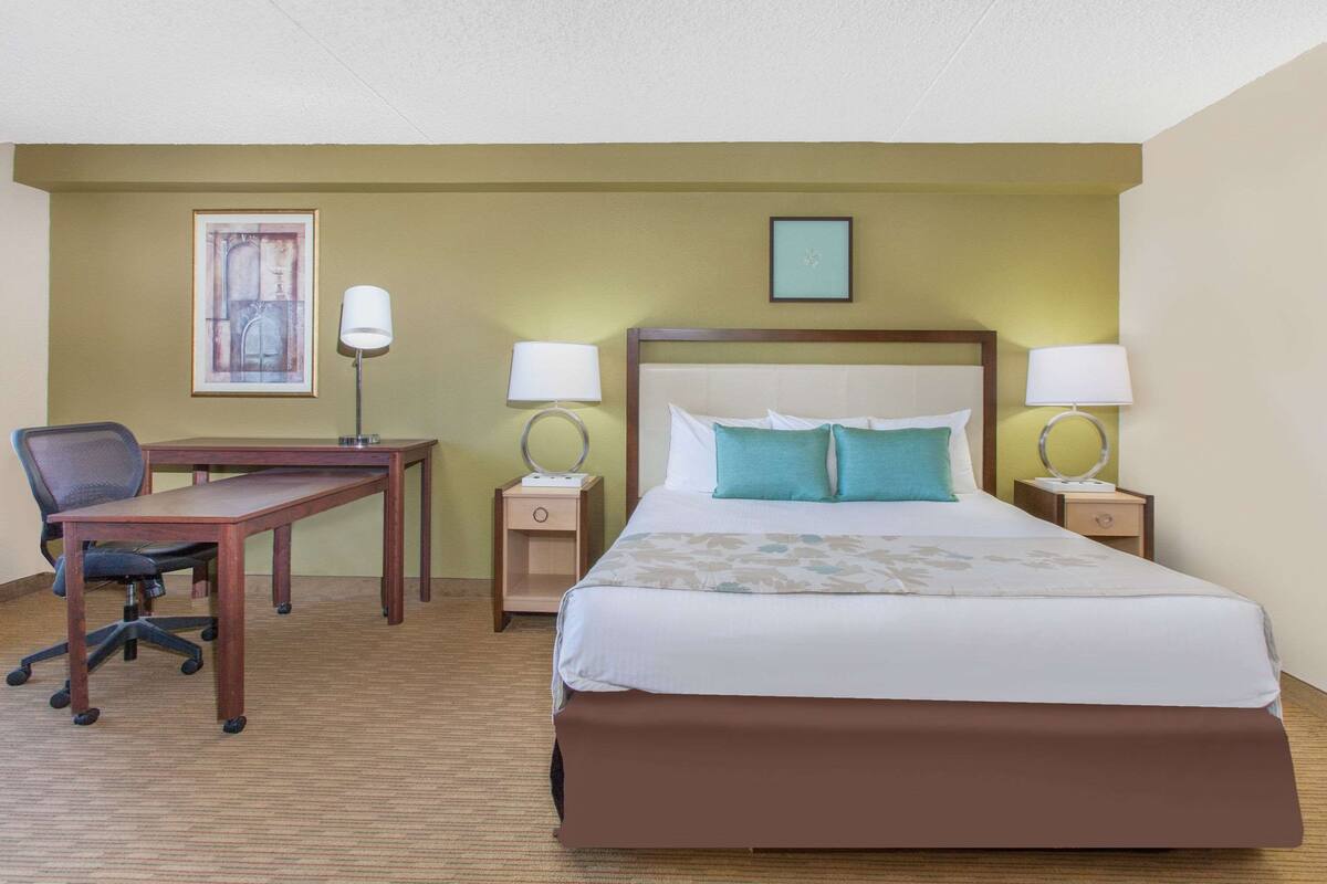 Ett hotellrum med en stor säng, ett skrivbord, två sänglampor, en stol och en inramad tavla på väggen.