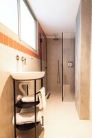 Un baño moderno con un lavamanos blanco, una ducha con cerramiento de vidrio y una ventana que permite la entrada de luz natural.