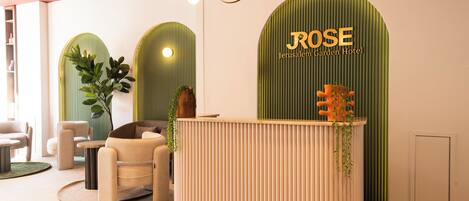A reception desk with a green wall and the name "ROSE Jerusalem Garden Hotel" above it. There are two chairs and a potted plant in the foreground.