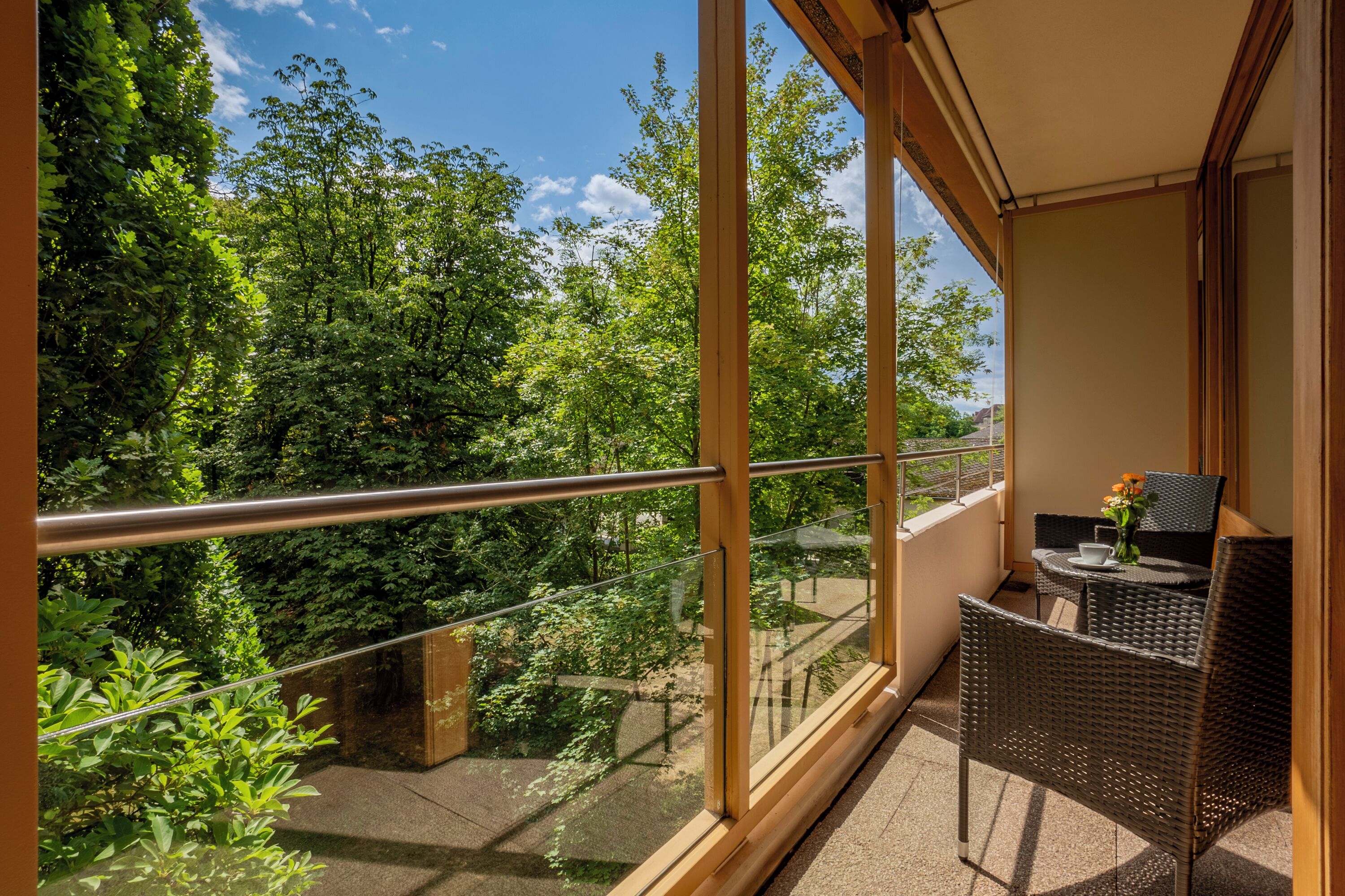 Ein Balkon mit Glastrennwand, Korbmöbeln und Blick auf üppiges Grün.