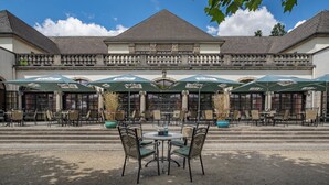 Un café en plein air avec des tables et des chaises, des parasols, et un bâtiment avec des colonnes et un balcon en arrière-plan.