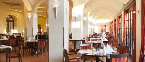 A spacious dining area with high ceilings and arched windows, featuring multiple tables and chairs arranged for dining.