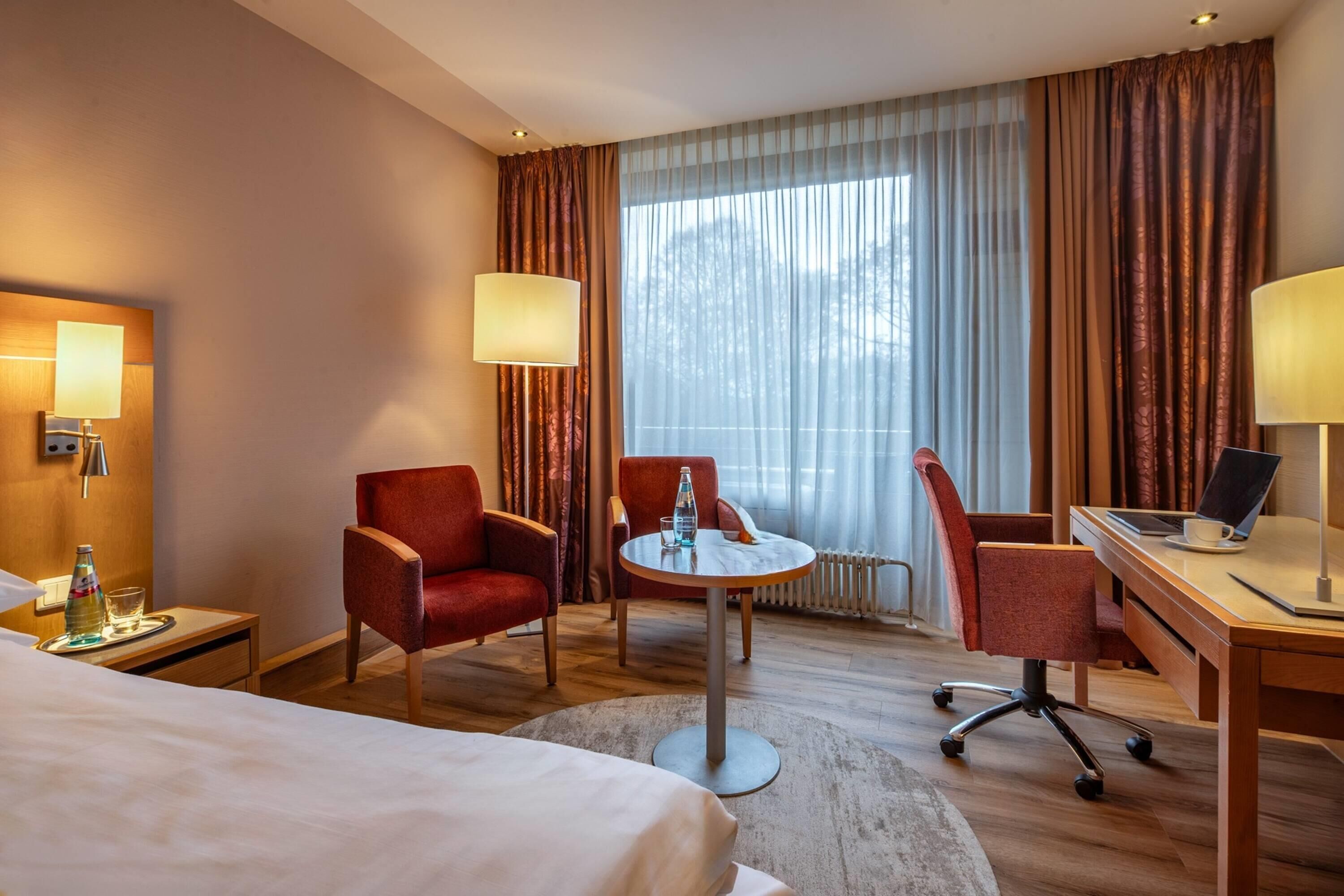 Quarto de hotel com uma cama, duas poltronas vermelhas, uma mesa redonda com abajur, uma escrivaninha com um laptop e uma janela grande com cortinas.