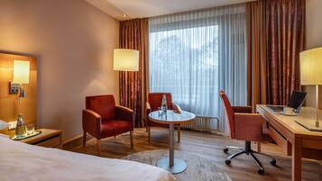 Quarto de hotel com uma cama, duas poltronas vermelhas, uma mesa redonda com abajur, uma escrivaninha com um laptop e uma janela grande com cortinas.
