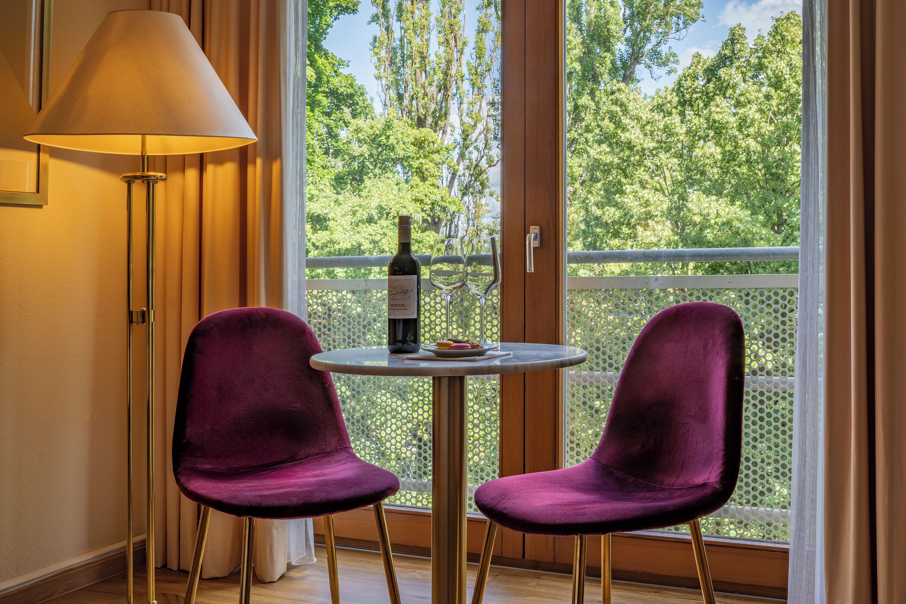 A room with a table, two chairs, a lamp, a bottle of wine, and a view of trees through a window.