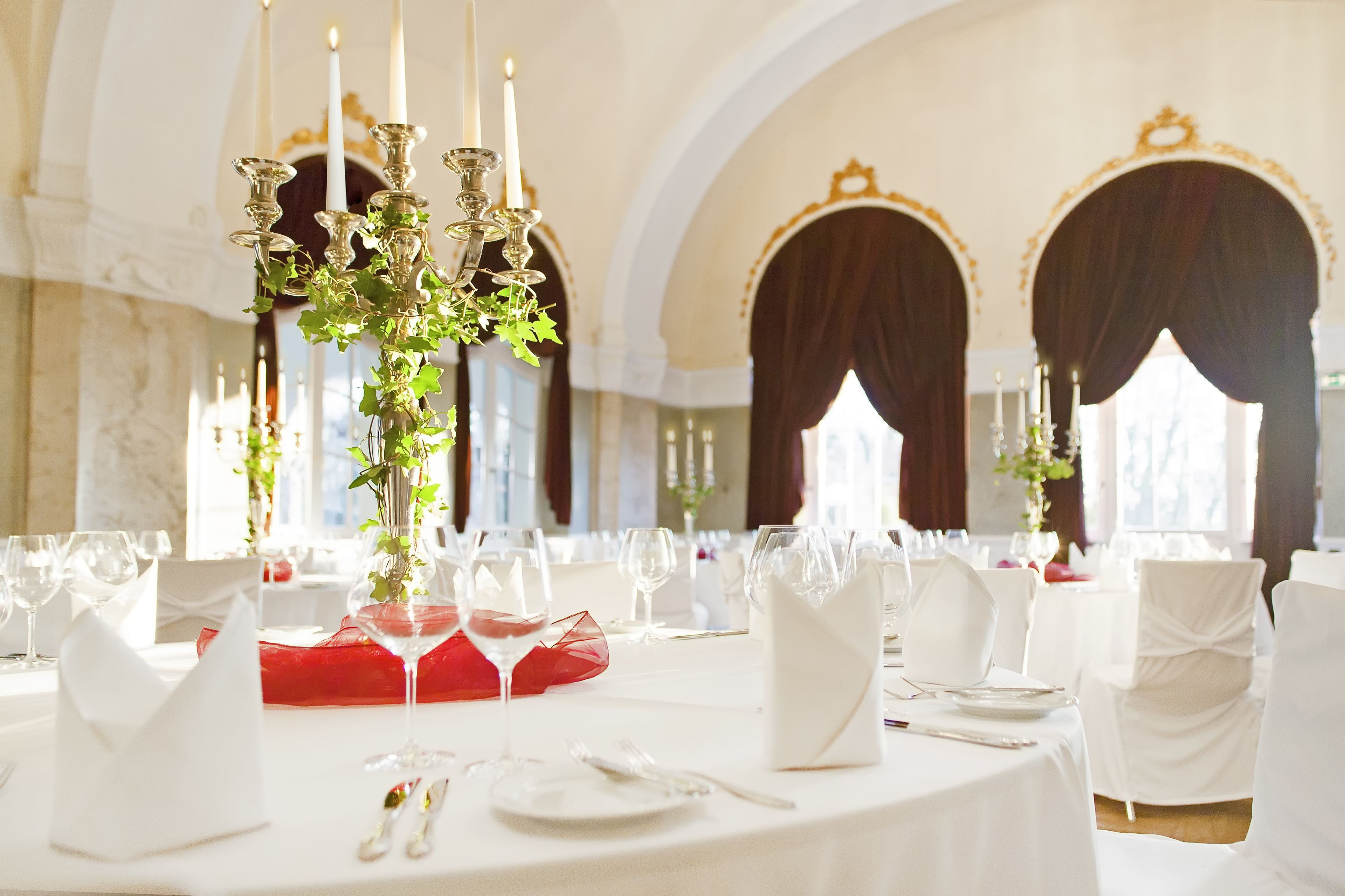 A banquet hall with round tables set for a formal event, featuring white tablecloths, red napkins, and elegant candelabras.